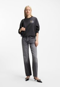 Ξανθιά γυναίκα που φοράει μαύρο φούτερ με μικρό λουλουδάτο σχέδιο, ξεθωριασμένο γκρι τζιν με κουμπιά, μαύρες μυτερές γόβες και κρατάει μαύρη τσάντα ώμου.