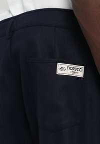 Artimiausiu planu matyti tamsiai mėlynų kelnių kišenę su balta stačiakampio formos Fiorucci Milano prekės ženklo etiketė, siuvinėta ant galinės juosmens dalies.