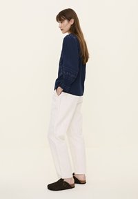 Marineblaue Bluse mit langen Ärmeln und gestickten floralen Mustern, kombiniert mit weißen, lockeren Hosen und dunklen Clogs mit Schnalle.