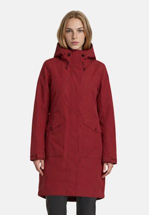 Junge Frau trägt eine lange rote Winterjacke mit Kapuze und Fronttaschen, steht vor einem schlichten hellen Hintergrund.