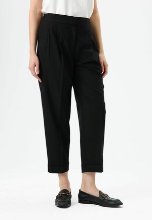 Pantalon noir sur mesure avec plis, longueur raccourcie, tissu lisse, assorti à des mocassins noirs ornés d'accents dorés. Design minimaliste.