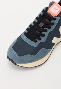 Sneaker blu navy con dettagli in suede azzurro chiaro, lacci neri e un'etichetta loggata rosa. Presenta un tessuto testurizzato e una suola di colore crema.