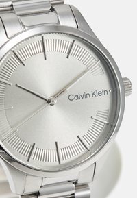 Reloj de acero inoxidable plateado con una esfera texturizada, marcadores de horas, agujas suaves y el logotipo de "Calvin Klein". Diseño de caja circular.