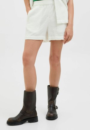 Shorts blancs texturés avec une taille élastique, associés à des bottes en cuir marron montantes avec des accents de boucles et une semelle robuste.