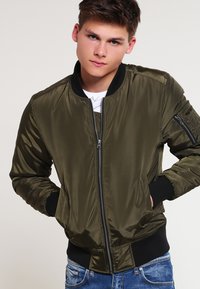 Olivovo zelená bomber bunda vyrobená z lesklého materiálu, s komfortným zipsom, elastickými manžetami a bočnými vreckami; vystavená nad bielym tričkom.