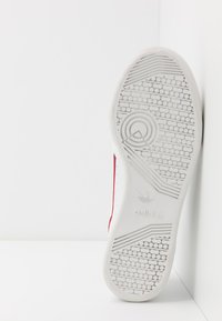 Fehér sneaker talpa texturált gumiból készült, mintázott futófelülettel. Piros díszítések láthatók az oldalakon. A márka logója a saroknál található.