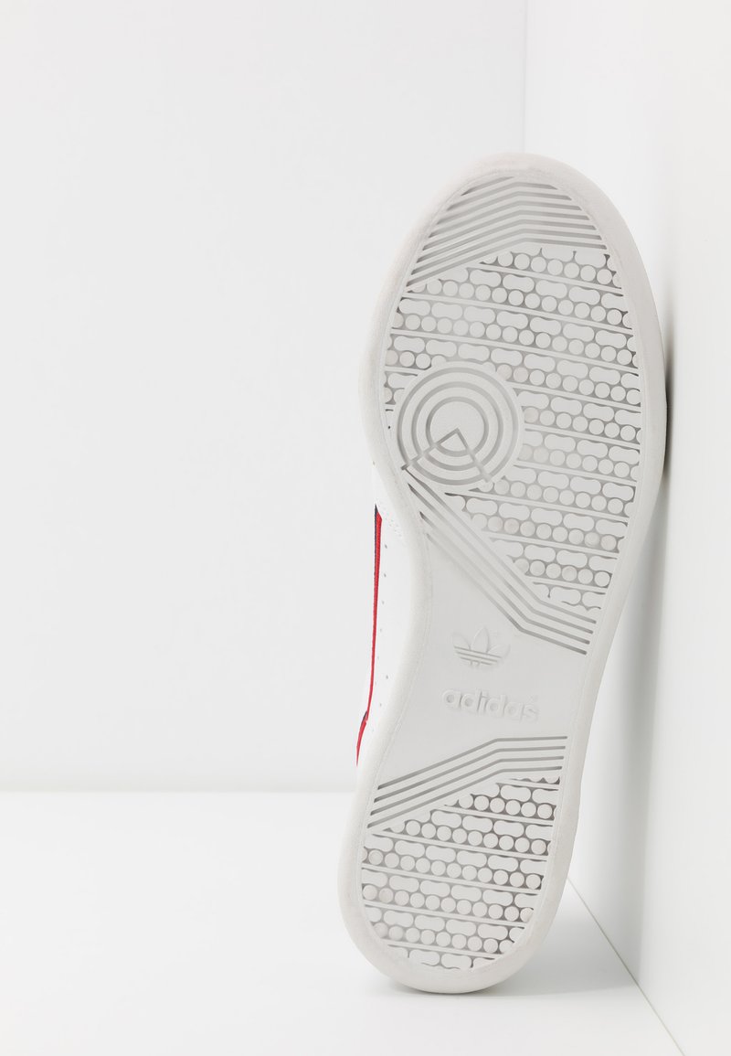 White sneaker outsole with textured rubber, featuring a patterned tread. Red accents visible on the sides. Brand logo present on heel.