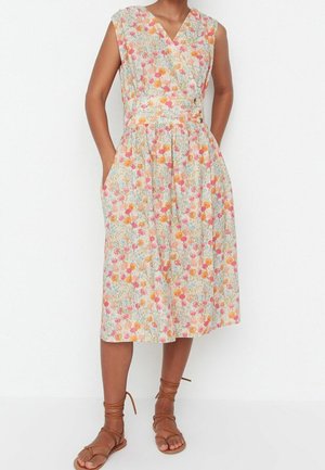 Frau trägt ärmelloses Midi-Kleid mit V-Ausschnitt und floralem Muster in Pink, Orange und Gelb sowie beige flache Sandalen mit Schnürung.