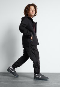 Chaqueta negra con capucha y bolsillos frontales, combinada con pantalones negros texturizados y zapatillas Nike grises. El estilo presenta un diseño contorneado y una tela texturizada.