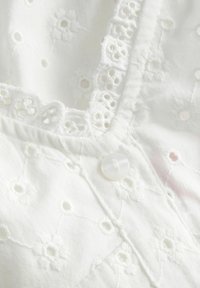 Tissu blanc avec broderie œillet florale, bordure en dentelle festonnée et un petit bouton rond blanc sur l'encolure d'un vêtement.