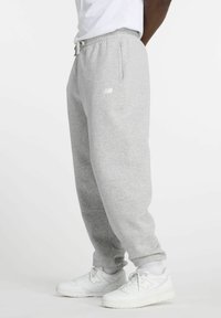 Šoninis vaizdas asmens, kuris dėvi pilkas New Balance sportines kelnes, baltus New Balance sportbačius, baltas kojines ir baltą marškinėlį, stovinčio ant vientiso fono.
