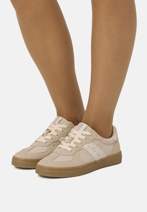 Zapatillas beige con cordones y suelas de goma, usadas con piernas desnudas, con rayas laterales blancas y etiquetas de marca en la lengüeta.