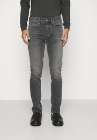 Vaqueros de mezclilla gris de corte ajustado, con cinco bolsillos, cierre de cremallera y detalles de costuras en contraste. Combinados con zapatos negros y una camiseta oscura.