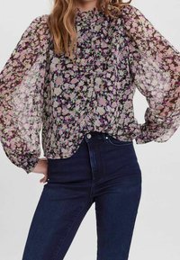 Höhytön kukkakuosinen pusero, jossa on pitkät, puhditut hihat ja röyhelökaulus. Moniväriset kukkakuviot tummalla taustalla. Yhdistetty tummiin farkkuihin.