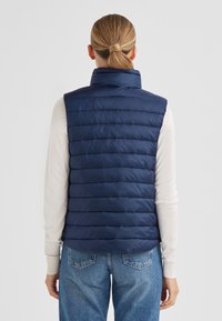 Marineblaue gesteppte Weste mit hohem Kragen, Steppmuster und Reißverschluss; getragen über einem hellfarbenen Langarmshirt und Jeans.