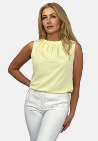 Krótki rękaw żółtej bluzki z plisowanym dekoltem, zestawionej z dopasowanymi białymi spodniami. Gładki materiał, minimalistyczny design, odpowiednie na co dzień lub na formalne okazje.