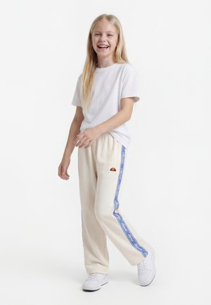 Uśmiechnięta dziewczyna z długimi blond włosami, ubrana w białą koszulkę, kremowe spodnie dresowe z niebieskimi paskami oraz białe trampki, stojąca na białym tle.