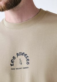 Beige ¾-ærmet t-shirt lavet af ribbet stof med et sort grafisk design, der står "LOS ANGELES" og "THE VELVET EMBER."