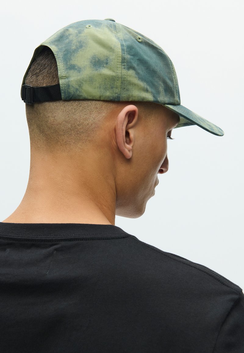 Green and blue tie-dye baseball cap features a curved brim and a black adjustable strap at the back. Worn with a black shirt.