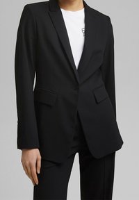 Czarna marynarka szyta na miarę z pojedynczym guzikiem, klapami oraz przednimi kieszeniami. Stylizowana na białym t-shircie, o gładkiej fakturze.