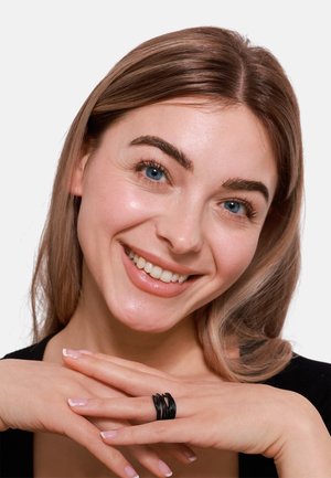 Anello multi-banda nero con bordi lisci e arrotondati, indossato su una mano con unghie curate. La modella mostra un'espressione amichevole.