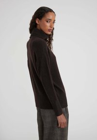 Vrouw met krullend haar, gekleed in een donkerbruine coltrui en grijs geruite broek, die zijwaarts staat tegen een effen lichte achtergrond.