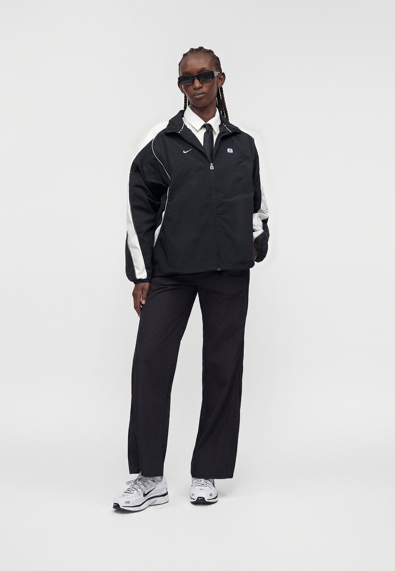 Persoon draagt een zwart-witte Nike jas, zwarte broek, witte sneakers, zwarte stropdas en zonnebril, staande tegen een effen lichte achtergrond.