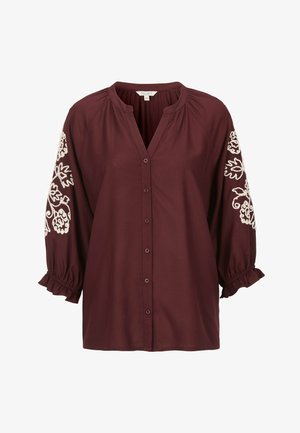Burgundisk blus med V-ringning, framknappar och 3/4 puffärmar som har vit blommig broderi och rynkade manschetter. Slät textur.