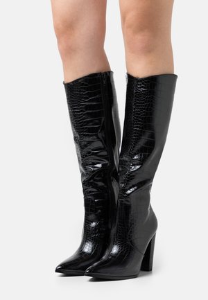 Woman wearing knee-high black crocodile-patterned leather boots with pointed toes and block heels on a white background.