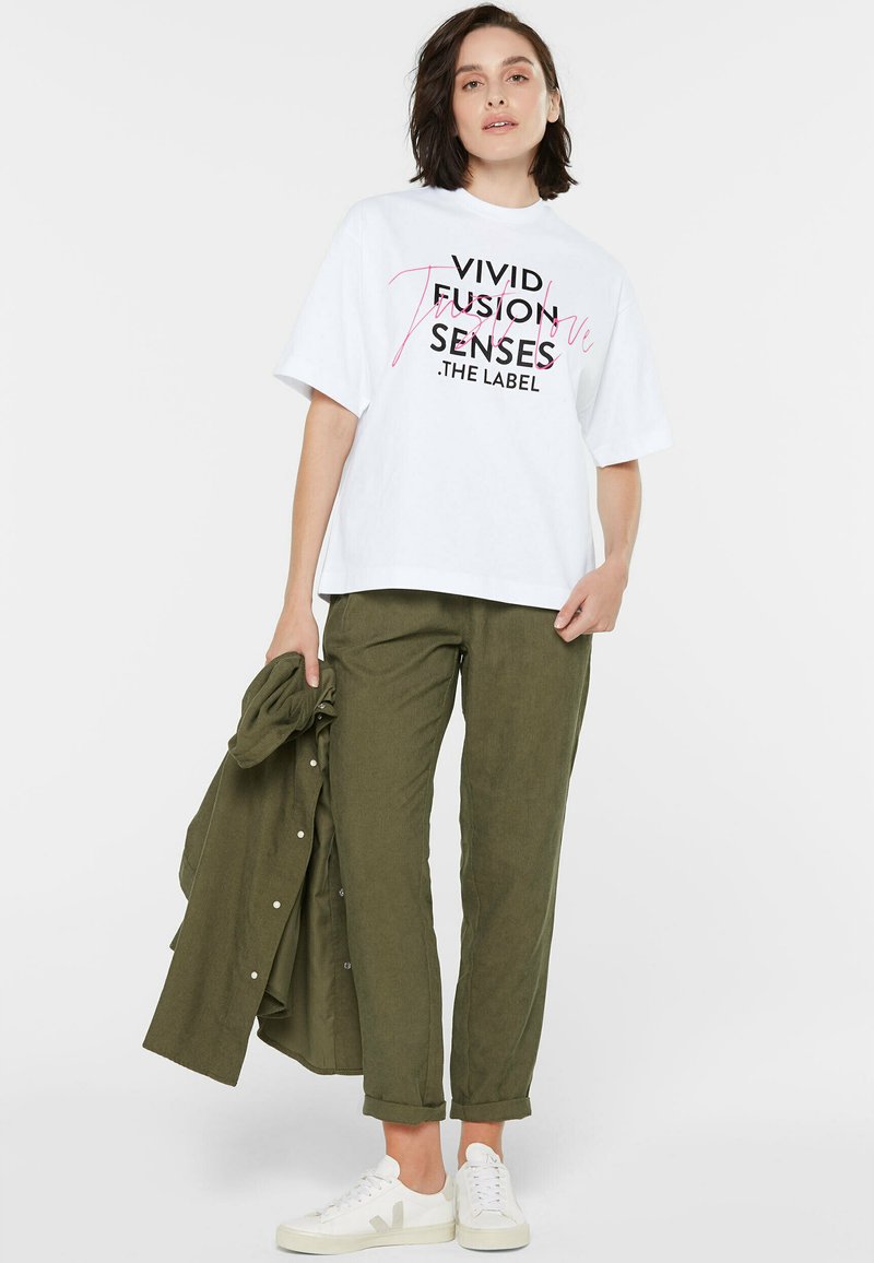 Weißes T-Shirt mit kurzen Ärmeln, bedruckt mit schwarzem und pinkem Text, kombiniert mit olivgrünen Hosen und weißen Sneakers. Das Outfit wird durch eine passende grüne Jacke ergänzt.