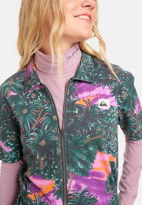 Kortermet jakke med glidelås i mørkegrønn med tropisk palmetrykk i rosa og lilla toner. Båret over en lys lilla topp med høy hals.