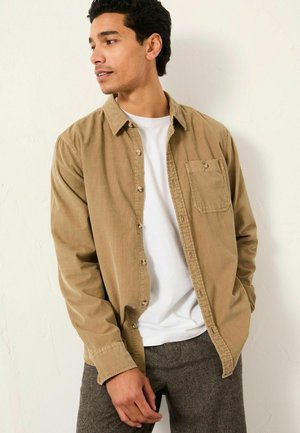 Beige Hemd mit Knöpfen aus weichem Stoff, mit einer Brusttasche, Kragen und langen Ärmeln, getragen über einem weißen T-Shirt.