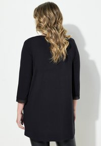 Zwarte tuniek met driekwart mouwen, voorzien van geaccentueerde textuur bij de manchetten en een rechte zoom. Gemaakt van zachte, lichte stof.