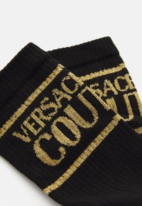 Čierne ponožky Versace Couture s zlatým trblietavým textom a pruhmi, rebrovaným manžetovým okrajom, preložené vedľa seba na bielom pozadí.