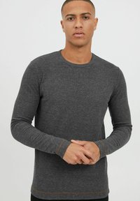 Langärmliges graues Shirt mit Rundhalsausschnitt, aus strukturiertem Stoff. Kontrastierende braune Nähte an den Bündchen und dem Saum. Minimalistisches Design.