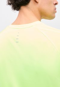 Mann trägt ein blassgelbes Sportshirt, zeigt den Nackenausschnitt hinten und kurze lockige Haare, seitlich vor einem einfarbigen Hintergrund.