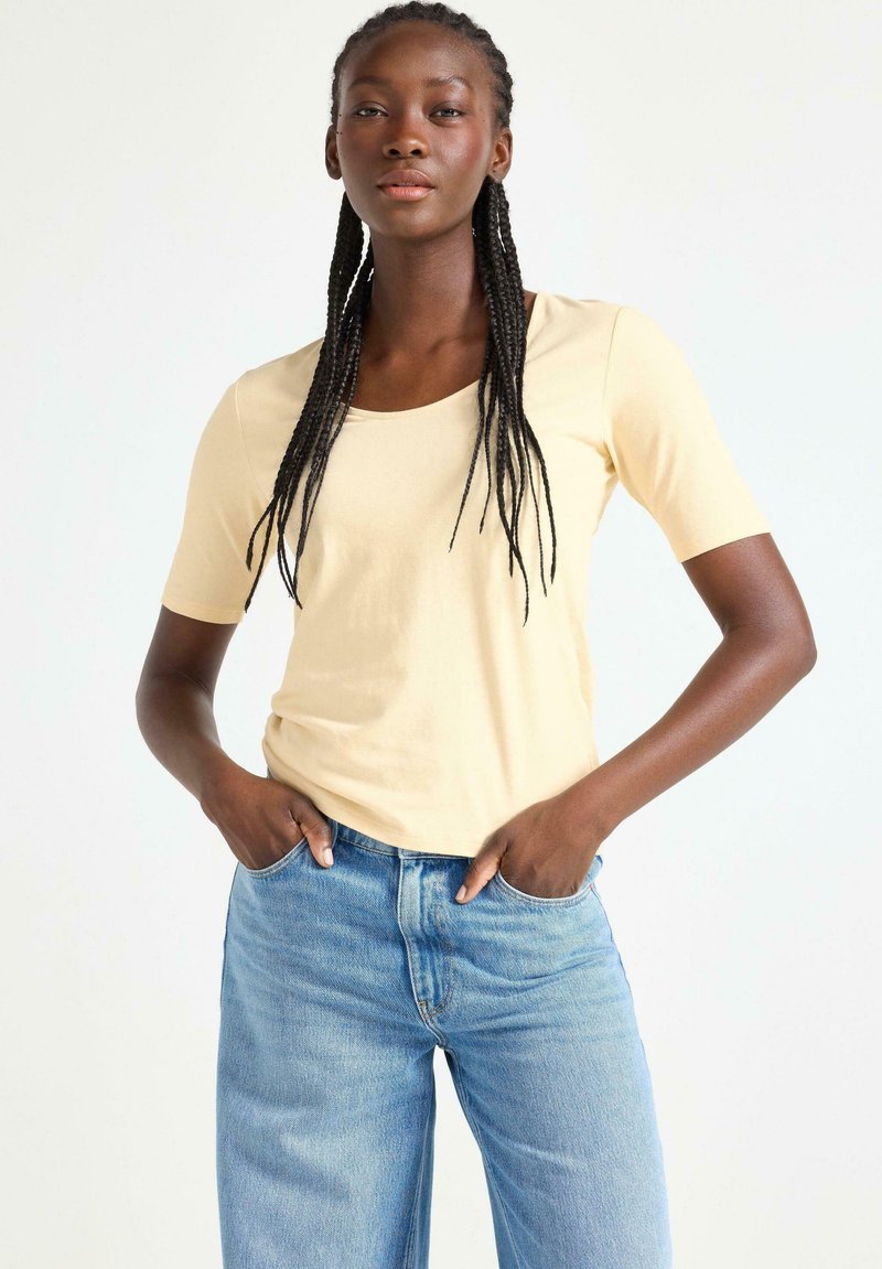 Kortærmet t-shirt med scoop-udskæring i lysegul bomuld, parret med lyseblå denim jeans. Modellen poser med hænderne i lommerne.