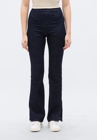 Φαρδιά πα ντελόνια από σκούρο ντένιμ με ψηλή μέση, που διαθέτουν λεία υφή και πλευρικές χαραγές στο τελείωμα, σε συνδυασμό με λευκά αθλητικά παπούτσια.