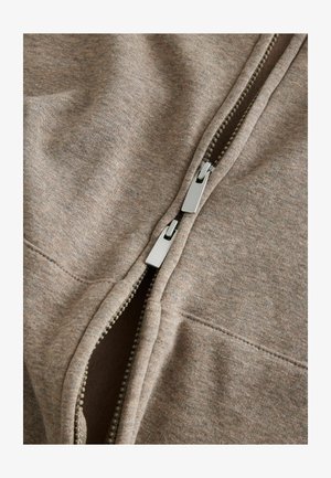 Világosbarna cipzáras ruhadarab, puha, texturált anyagból. A cipzár ezüst színű és középen helyezkedik el.