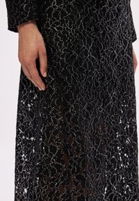 Juoda nėriniuota suknelė su sidabriniais gėlių raštais, išsiskirianti tekstūruotu paviršiumi. Ranką švelniai atsirėmus į audinį, paryškintas detalumas.