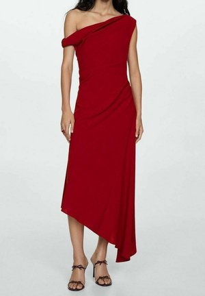 Kobieta ubrana w czerwoną, asymetryczną sukienkę midi na jedno ramię oraz czarne, wysokie sandały na paski, stojąca na jednolitym tle.