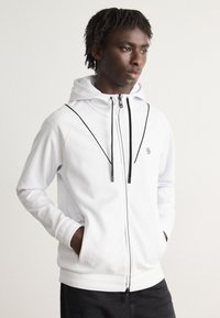 Veste à capuche blanche avec capuche, accents noirs, deux poches latérales et un logo sur la poitrine. Texture lisse et éléments de design moderne.