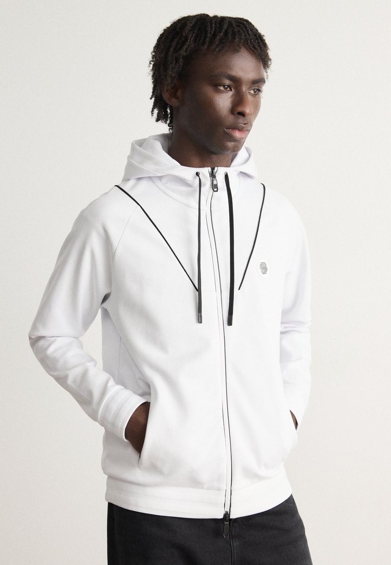 Veste à capuche blanche avec capuche, accents noirs, deux poches latérales et un logo sur la poitrine. Texture lisse et éléments de design moderne.