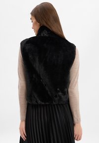 Czarna kamizelka z futra syntetycznego z wysokim kołnierzem, noszona na beżowej bluzce z długim rękawem. Posiada krótki fason, gładką teksturę i brak widocznych zapięć.