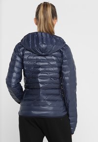 Doudoune bleu marine à capuche, dotée d'un design matelassé, d'une texture brillante et d'une silhouette ajustée. Fabriquée en tissu léger et isolant.