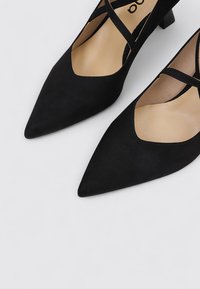 Zapatos de ante negros de punta afilada con un tacón bajo y correas en la ankle entrecruzadas. Textura suave y diseño minimalista.