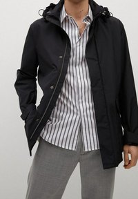 Musta ulkotakki hupulla, jossa on vetoketju ja painonappisuljenta, puettuna raidallisen paidan ja harmaiden housujen päälle. Sileä pinta ja rento istuvuus.