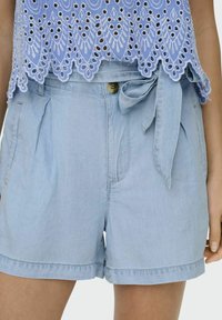Calções de ganga de cor azul clara com um laço na cintura, apresentando pregas e bolsos laterais. O tecido tem uma textura suave com beiras de bainha dobradas.