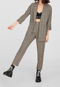 Costume à carreaux marron et noir avec un blazer court et un pantalon ajusté. Porté sur un top en dentelle noire, assorti à des bottes noires épaisses.