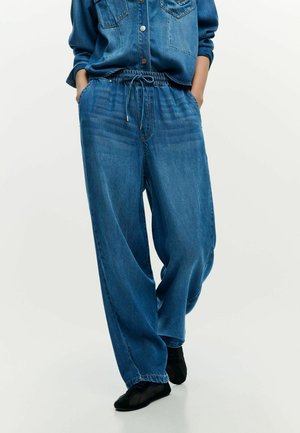 Inimene kannab lahtisi siniseid teksapükse, millel on elastne vöökoht ja pael, kombineerituna teksasärgiga ja mustade kingadega.